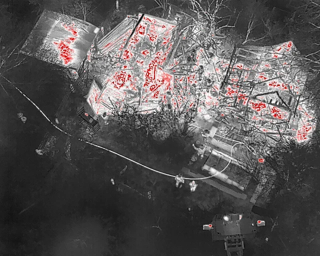 Eine Luftaufnahme in Thermalsicht zeigt ein brennendes Gebäude, umgeben von Feuerwehrleuten, die den Brand bekämpfen.