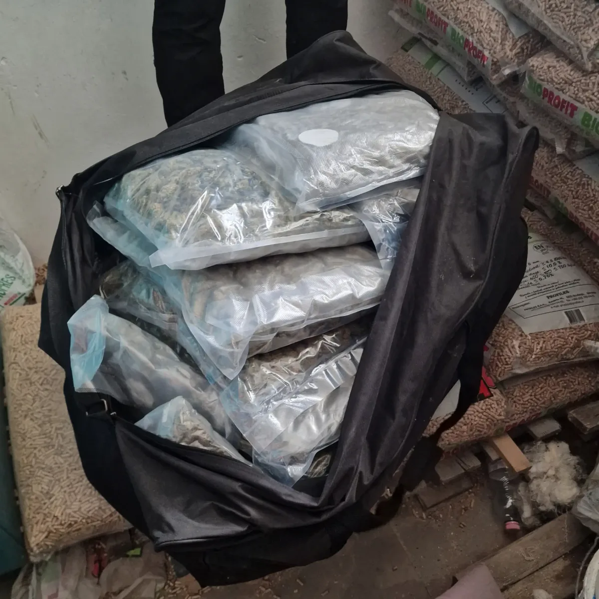 Ein schwarzer Rucksack liegt offen auf dem Boden und ist gefüllt mit mehreren durchsichtigen Plastikbeuteln, die Marihuana enthalten. Der Raum wirkt wie ein Lager mit weiteren Säcken im Hintergrund.