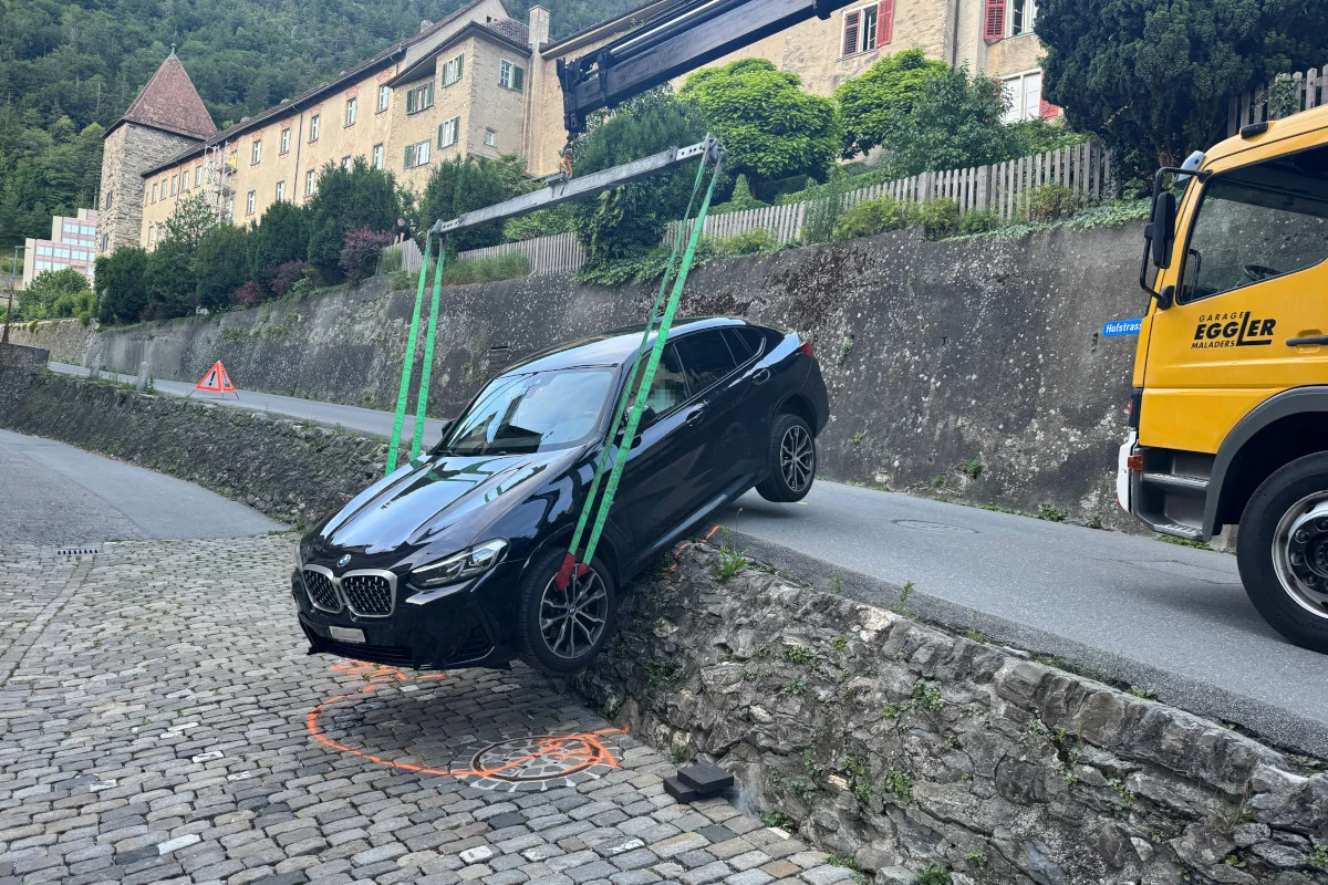 Ein schwarzes Auto hängt über einer Mauer und wird mit grünen Gurten von einem Kran gehalten. Ein gelber Lkw ist daneben zu sehen.