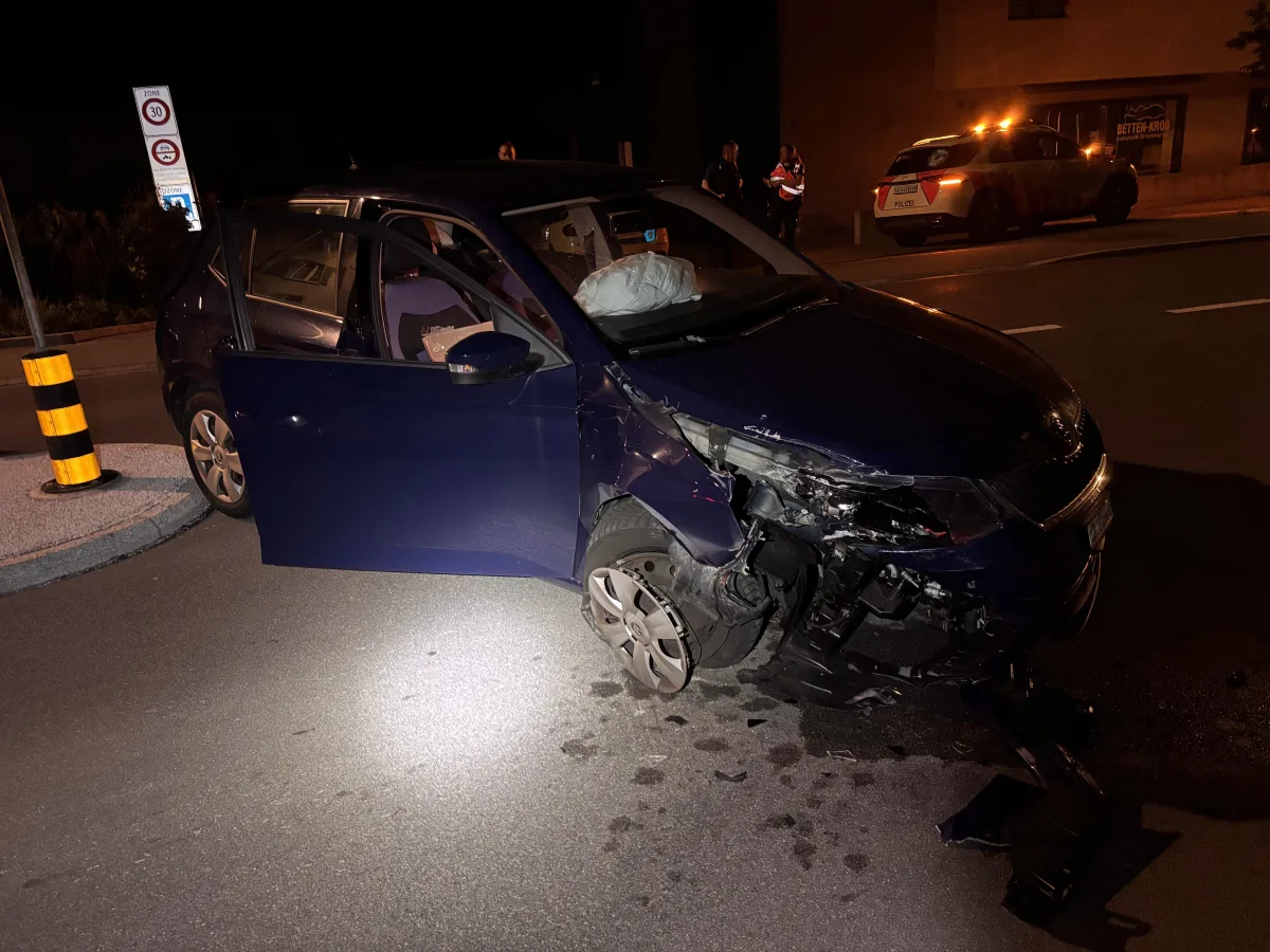 Ein blaues Auto mit erheblichen Frontschäden steht an einer dunklen Kreuzung. Ein Verkehrsschild ist ebenfalls beschädigt.