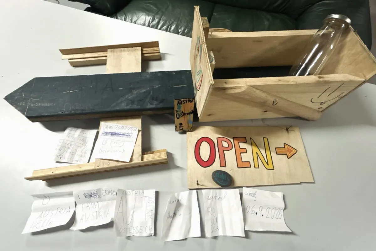 Eine Holzkiste liegt offen auf einem Tisch, umgeben von mehreren Zetteln und einer Glasflasche. Auf den Zetteln sind handgeschriebene Notizen zu sehen.