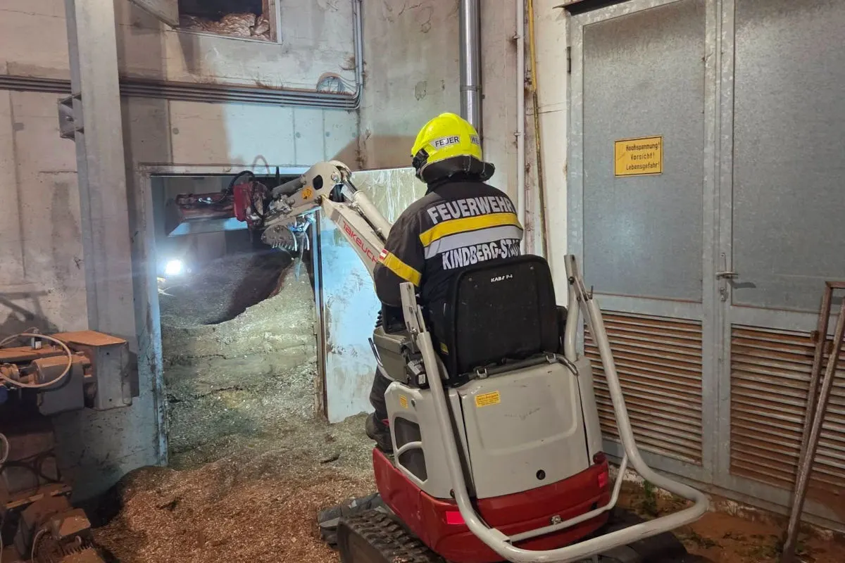 Ein Feuerwehrmann mit gelbem Helm sitzt auf einem kleinen Bagger in einem Gebäude. An der Wand ein Warnschild.