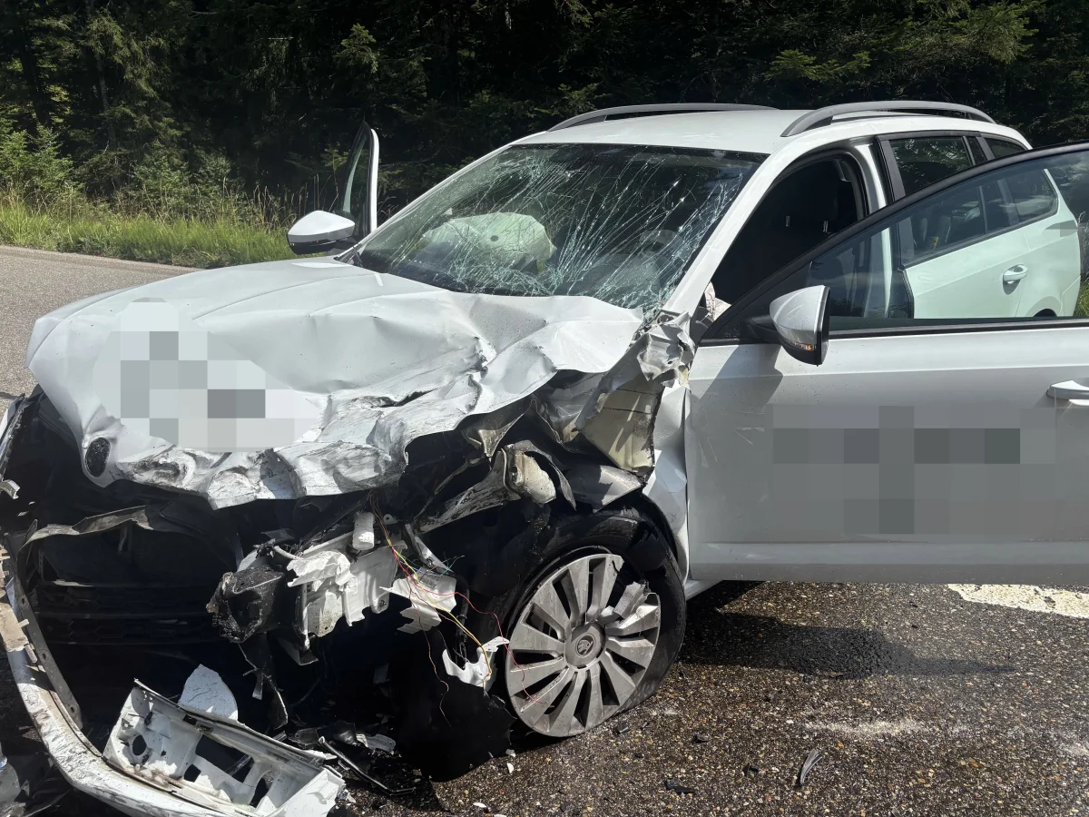 Ein weißes Auto mit schweren Frontschäden steht mit geöffneter Tür auf einer Straße. Der Unfall hat den Kotflügel und die Frontstoßstange stark verformt.
