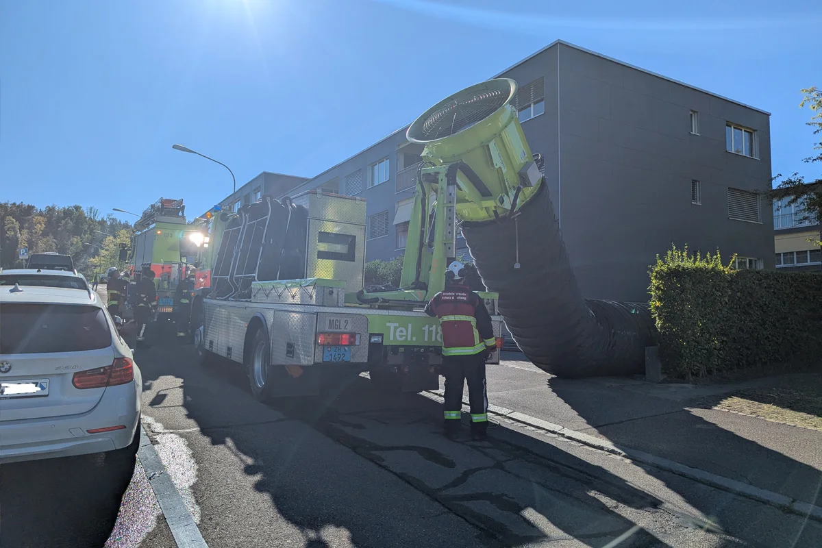 Ein Feuerwehrfahrzeug mit großem Belüftungsgerät ist vor einem Wohngebäude im Einsatz. Feuerwehrleute in Uniform befinden sich vor Ort.