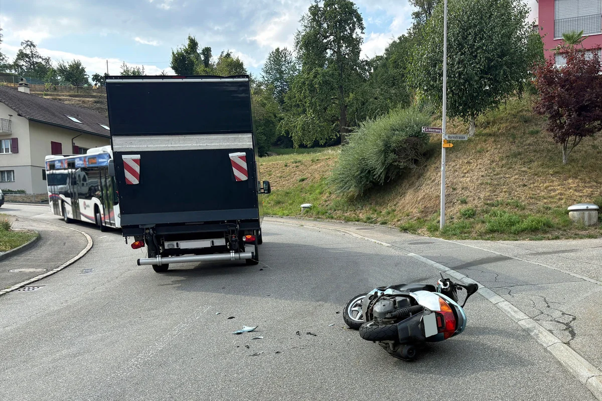 Ein umgestürzter Motorroller liegt auf einer asphaltierten Straße, neben einem Bus und einem Lkw im Hintergrund. Der Bereich ist grün bewachsen und wirkt städtisch.