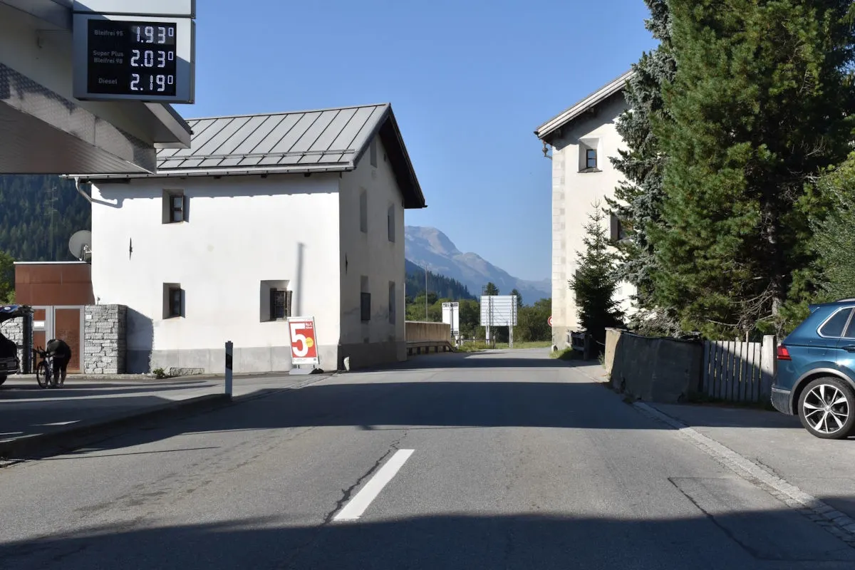 Eine leere Straße zwischen zwei weißen Gebäuden, mit einem Werbeschild an einer Tankstelle und einem Berg im Hintergrund.