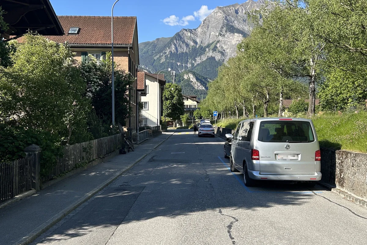 Ein silberner VW-Transporter steht rechts in einer ruhigen Straße geparkt. Im Hintergrund sind hohe Berge und Bäume zu sehen.