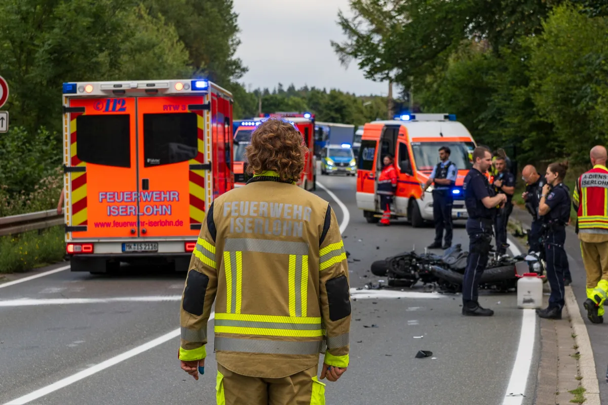 Ein Feuerwehrmann steht auf einer Straße; mehrere Einsatzfahrzeuge und ein umgestürztes Motorrad sind zu sehen. Einsatzkräfte sind vor Ort.