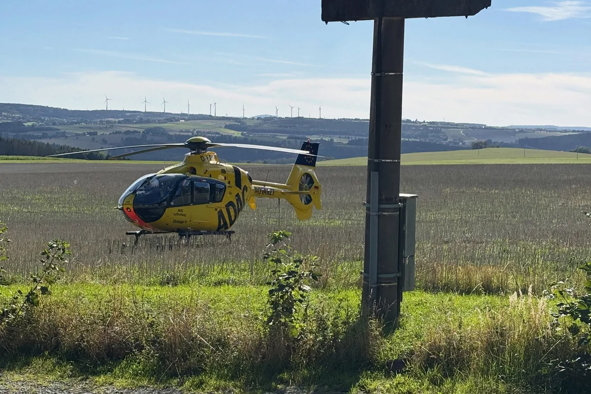 Ein gelber ADAC-Rettungshubschrauber steht in einem Feld. Im Hintergrund sind Hügel mit Windrädern zu sehen.