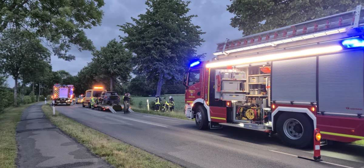 Zwei Feuerwehrfahrzeuge sind auf einer Landstraße im Einsatz. Einsatzkräfte stehen auf der Straße, während ein Auto auf einem Abschleppwagen verladen wird.