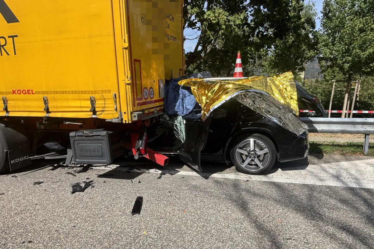 Ein schwarzes Auto ist schwer beschädigt und unter der Ladefläche eines gelben Lkw eingeklemmt. Der Unfall ereignete sich auf einer Straße mit Bäumen und einer Leitplanke.