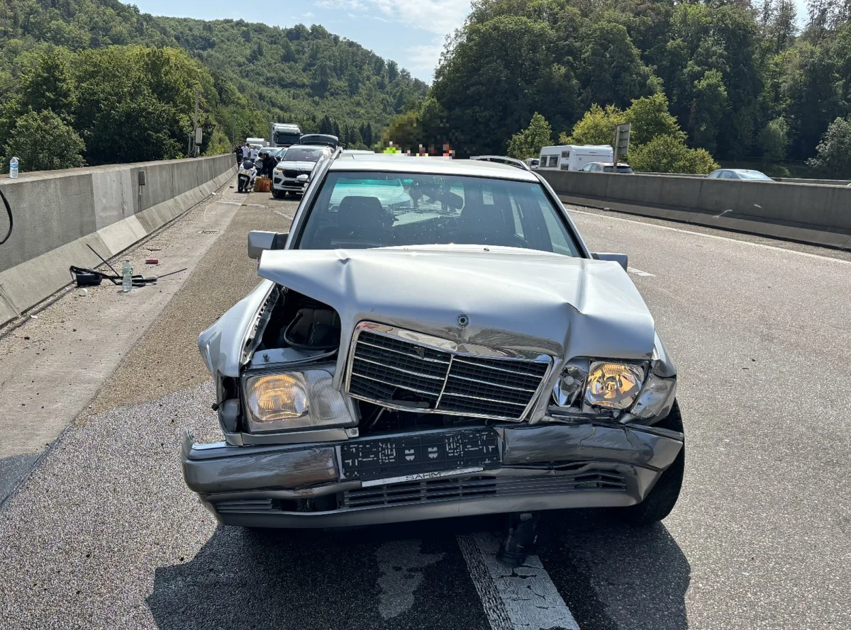 Ein silberner Mercedes-Benz mit stark beschädigter Front steht quer auf einer Autobahn. Im Hintergrund sind weitere Fahrzeuge und Bäume zu sehen.