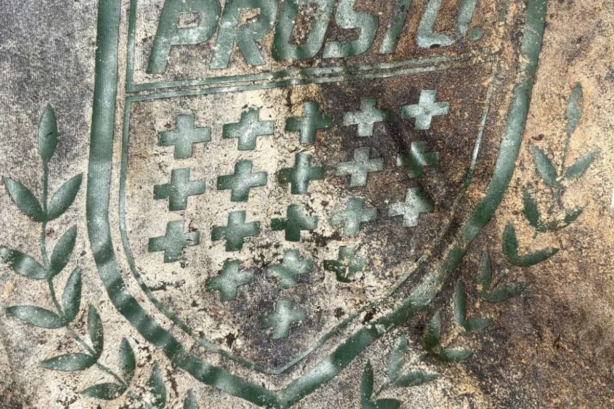 Detailliert graviertes Wappen im Boden mit dem Text 'PROST!' und mehreren kleinen Kreuzen umgeben von Lorbeerblättern.