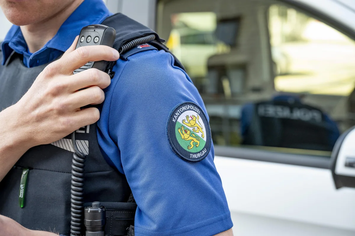 Ein Polizist der Kantonspolizei Thurgau hält ein Funkgerät vor einem Polizeifahrzeug. Das Emblem ist deutlich sichtbar.