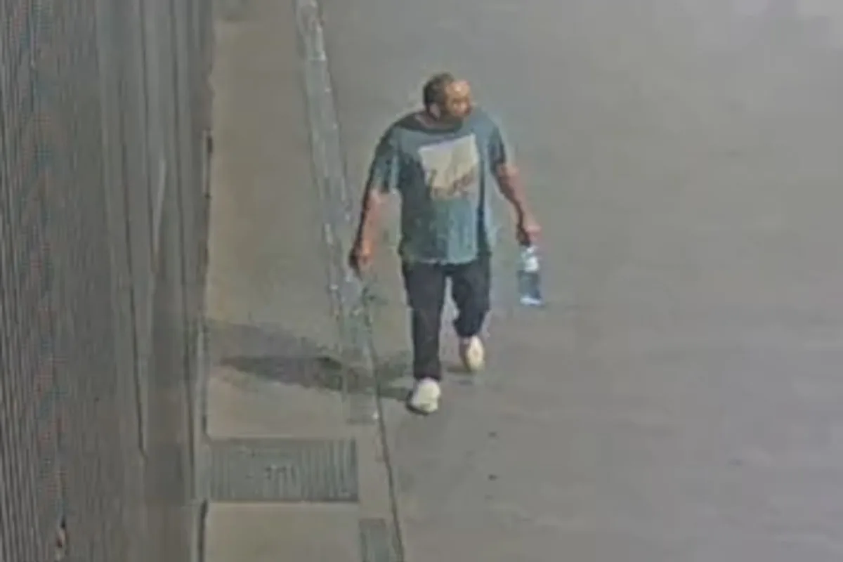 Ein Mann in dunkler Kleidung trägt eine Wasserflasche und geht entlang einer leeren Straße. Die Szene wirkt nächtlich.