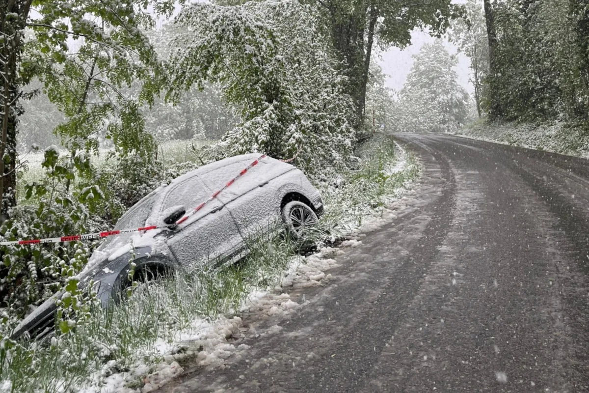 Ein Auto, das mit Schnee bedeckt ist, liegt in einem Graben neben einer verschneiten Landstraße. Bäume säumen den Weg.