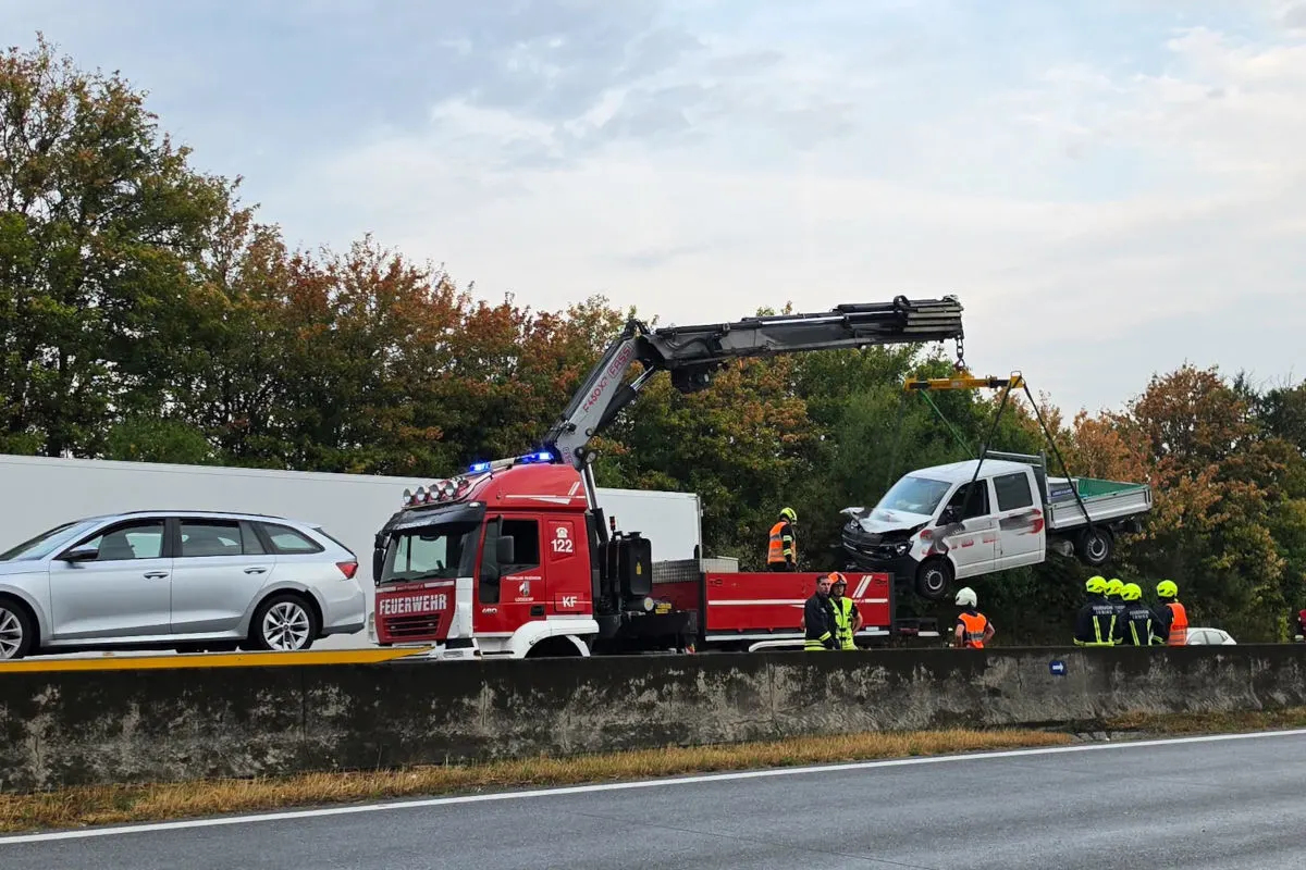 Ein Feuerwehrkran hebt ein beschädigtes Fahrzeug von der Autobahn an. Feuerwehrkräfte stehen daneben und beobachten den Vorgang.
