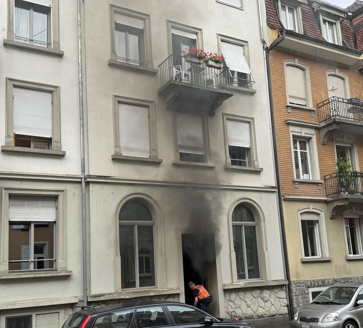 Dichter Rauch steigt aus der Eingangstür eines mehrstöckigen Wohnhauses auf. Ein Mann in oranger Weste steht in der Tür.