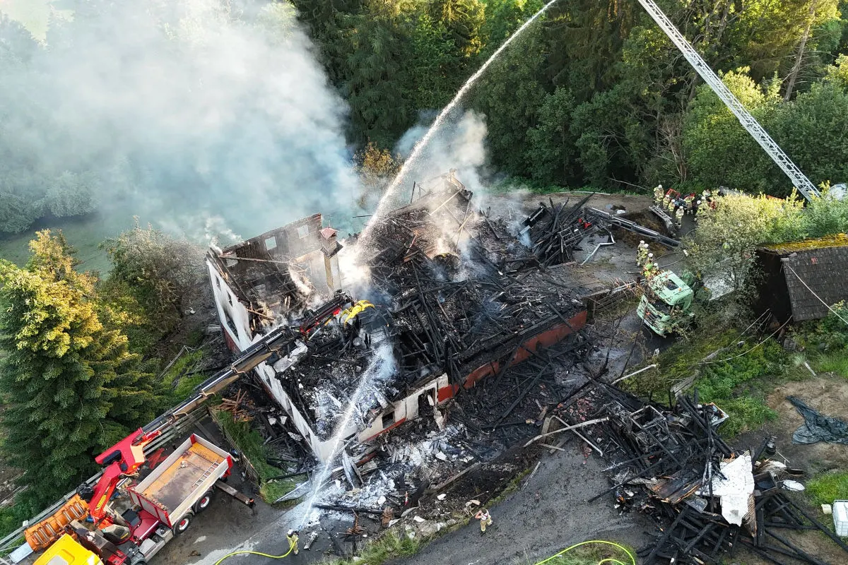Von oben betrachtet ist ein vollständig zerstörtes Gebäude zu sehen, umgeben von Rauch, während die Feuerwehr mit Löschmaßnahmen beschäftigt ist. Zwei Fahrzeuge, darunter eines der Feuerwehr, sind vor Ort.