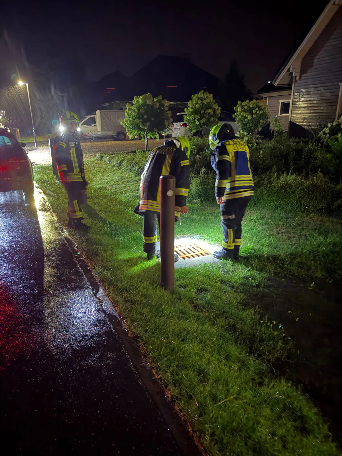 Drei Feuerwehrleute in Einsatzkleidung beugen sich bei Nacht über einen beleuchteten Gullydeckel auf einer nassen Straße.