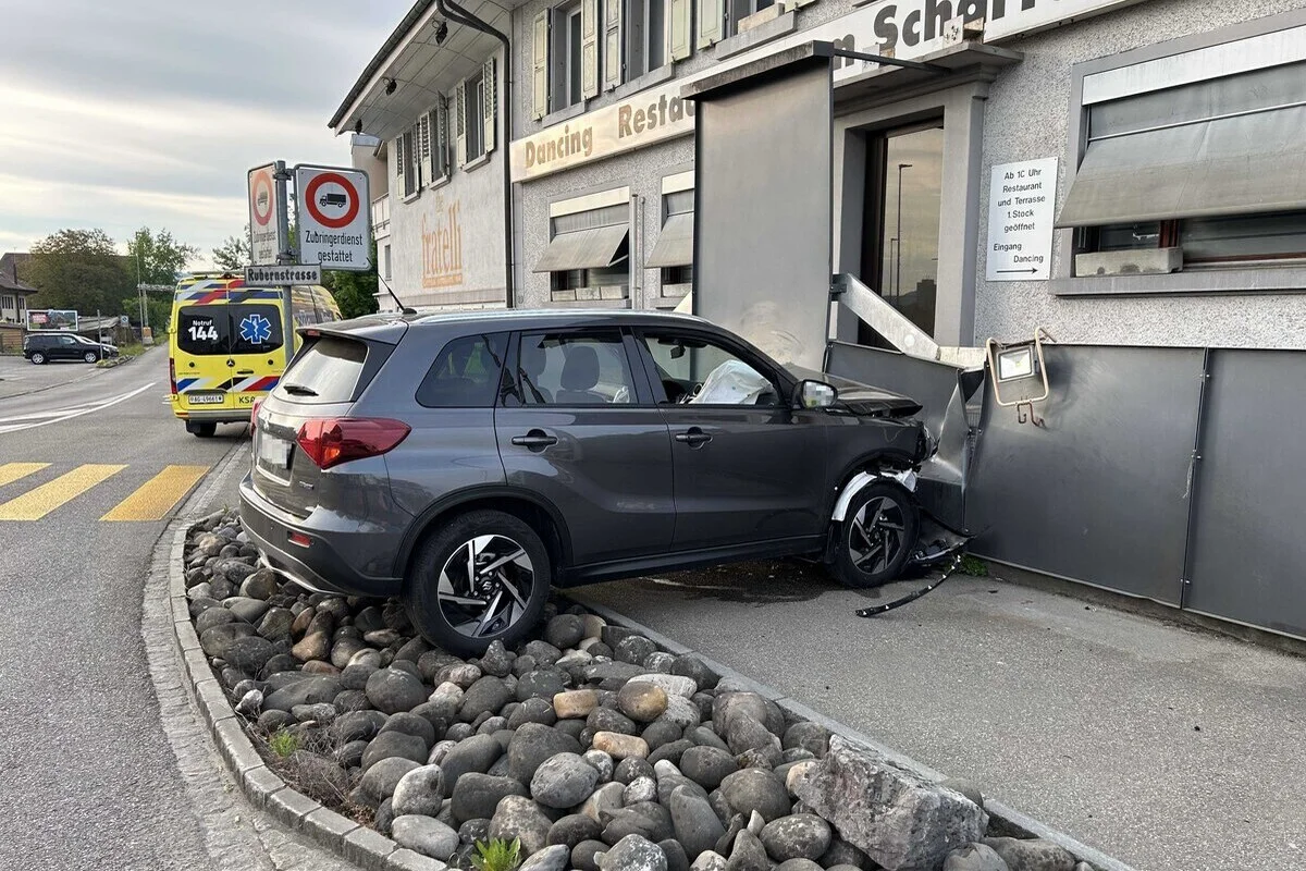 Ein graues Auto ist gegen die Wand eines Gebäudes gefahren, mit sichtbarem Schaden. Im Hintergrund ist ein Ambulanzfahrzeug erkennbar.