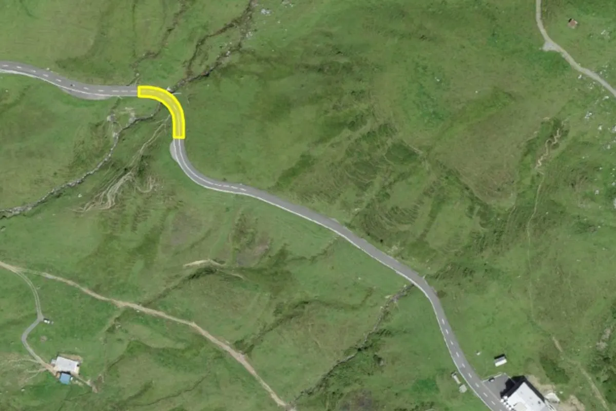 Die Luftaufnahme zeigt eine scharfe Kurve auf einer Landstraße in einer grünen, hügeligen Landschaft. Es sind keine Fahrzeuge oder Personen zu sehen, nur die Straße und umliegendes Grasland.