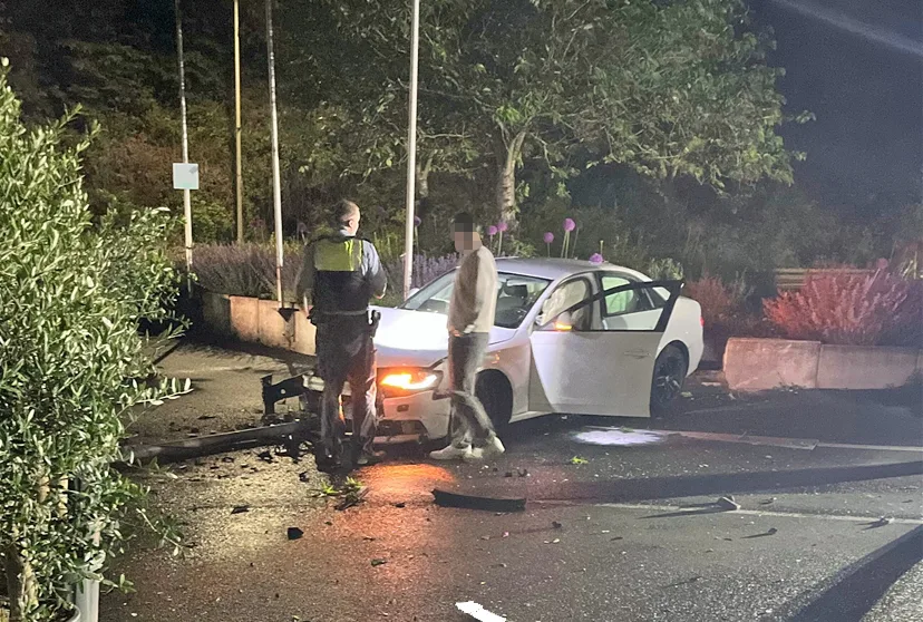 Ein weißes Auto steht nachts am Straßenrand, sichtbar beschädigt, mit offenem Fahrerfenster und zwei Personen daneben.