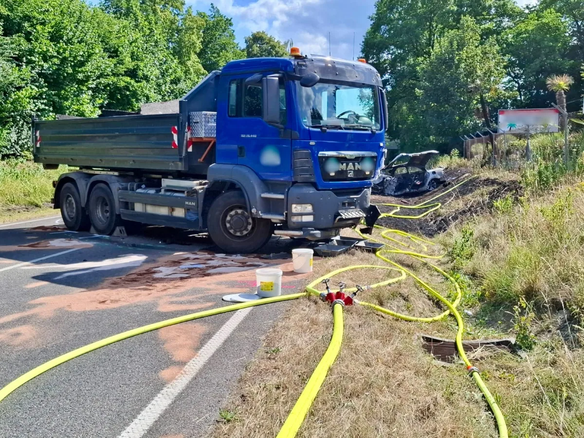 Auf einer Landstraße steht ein blauer MAN-Lastwagen. Rechts im Hintergrund befindet sich ein ausgebranntes Fahrzeugwrack.