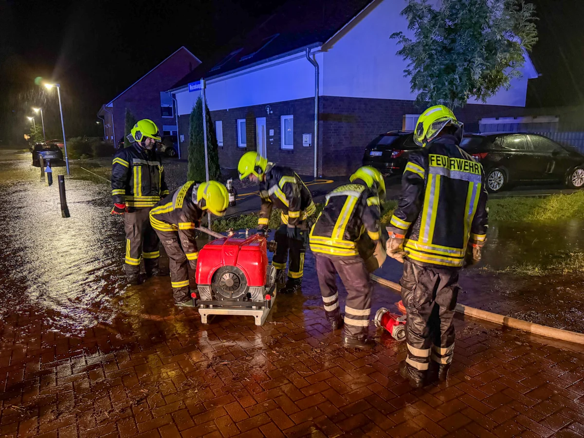 Mehrere Feuerwehrleute in Einsatzkleidung mit gelben Helmen arbeiten in einer regnerischen Nacht an einer roten Pumpe auf einer nassen Straße neben Wohnhäusern.