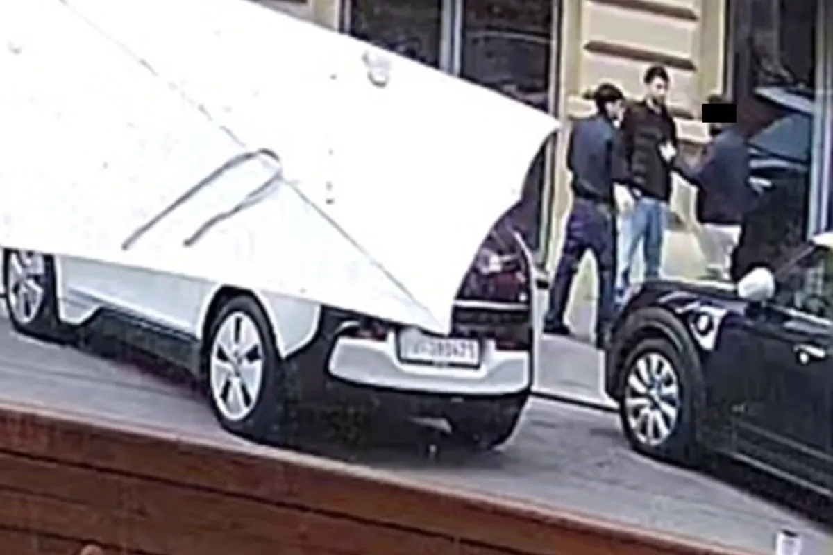 Zwei Personen in dunkler Kleidung stehen vor einem Gebäude. Im Vordergrund sind zwei geparkte Autos sichtbar.