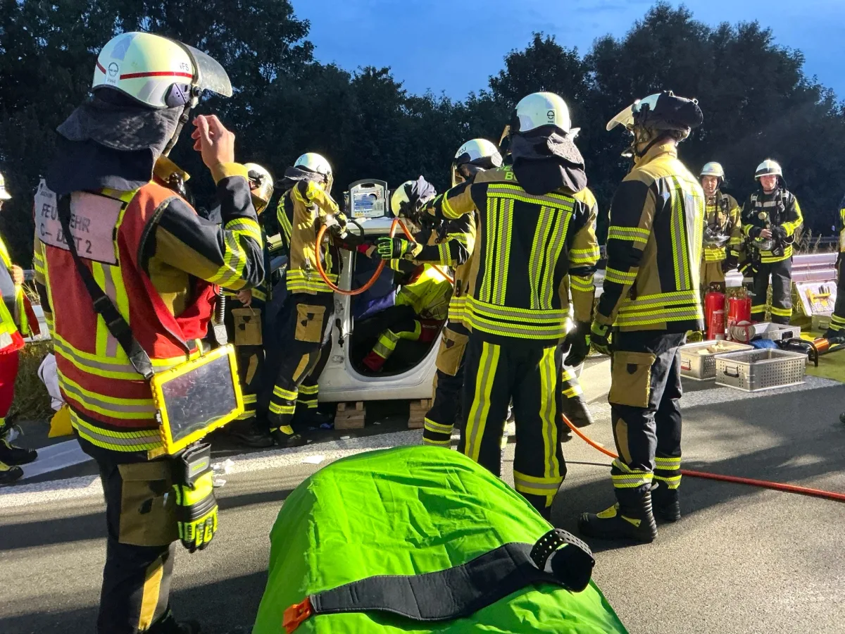 Eine Gruppe von Feuerwehrleuten in Schutzkleidung umgibt ein auf der Seite liegendes Auto bei Dämmerung. Verschiedene Werkzeuge und Ausrüstungen sind um sie herum zu sehen.