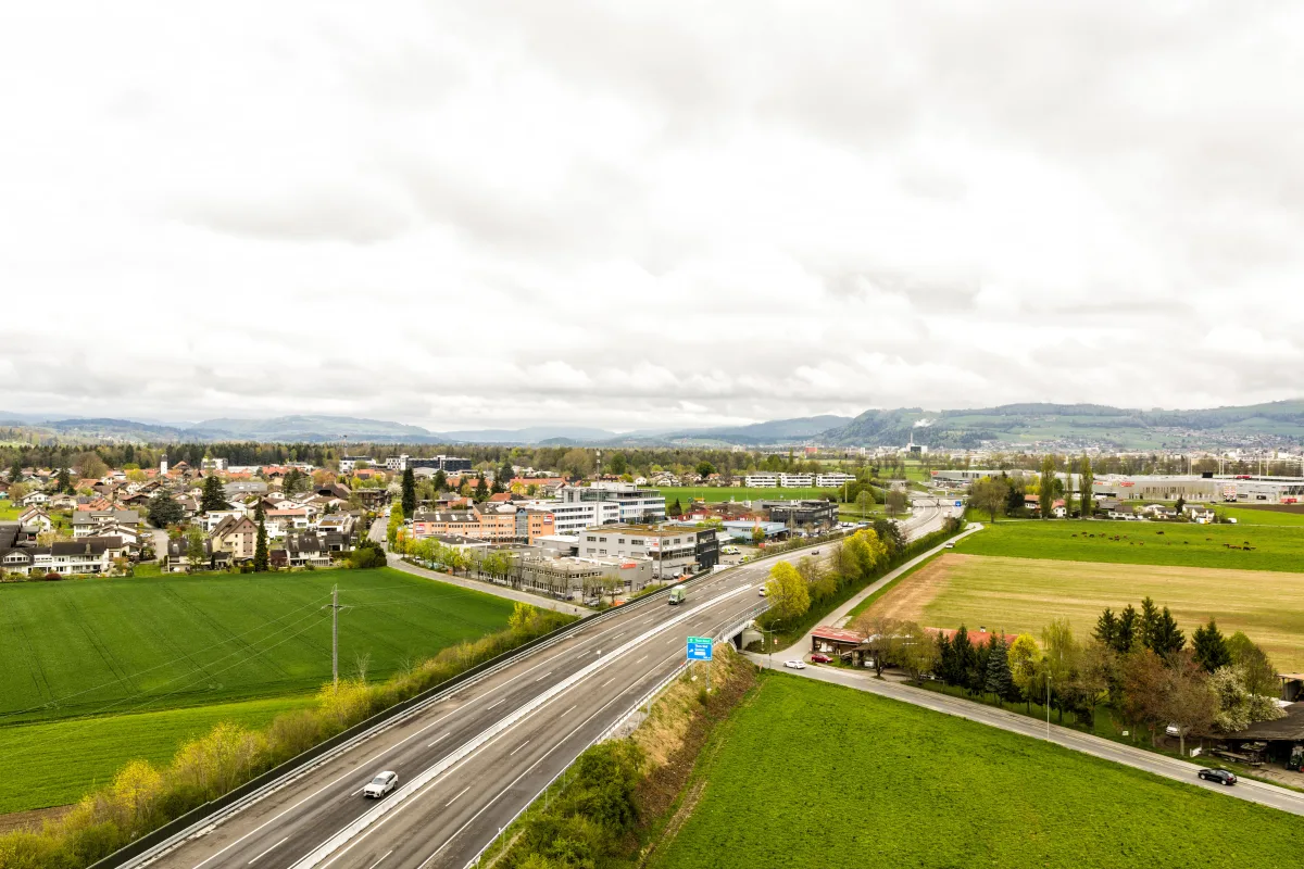 Eine zweispurige Autobahn verläuft durch eine ländliche Gegend, umgeben von grünen Feldern und vereinzelten Gebäuden unter bewölktem Himmel.