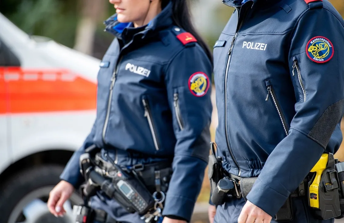 Zwei Polizisten in Uniform der Kantonspolizei Zürich stehen vor einem weißen Polizeifahrzeug mit orangefarbenem Streifen.