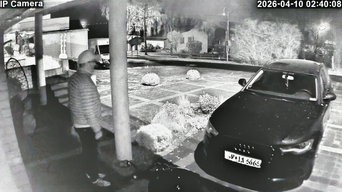 Eine Überwachungskamera erfasst eine Person, die neben einem schwarzen Auto steht, in einem beleuchteten Außenbereich bei Nacht.