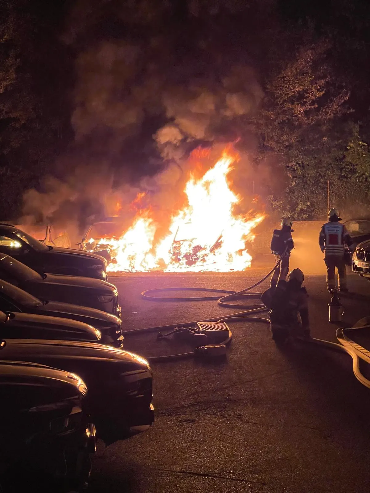 Ein Auto steht in Flammen auf einem Parkplatz bei Nacht. Feuerwehrleute sind mit Löschschläuchen im Einsatz, um das Feuer zu bekämpfen.