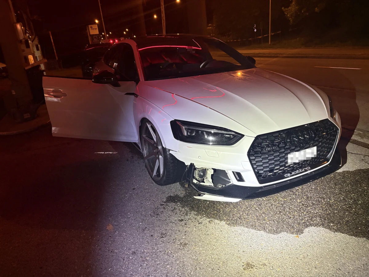 Ein weißer Audi steht bei Nacht mit geöffneter Fahrertür und beschädigter Front auf einer Straße an einer Tankstelle.