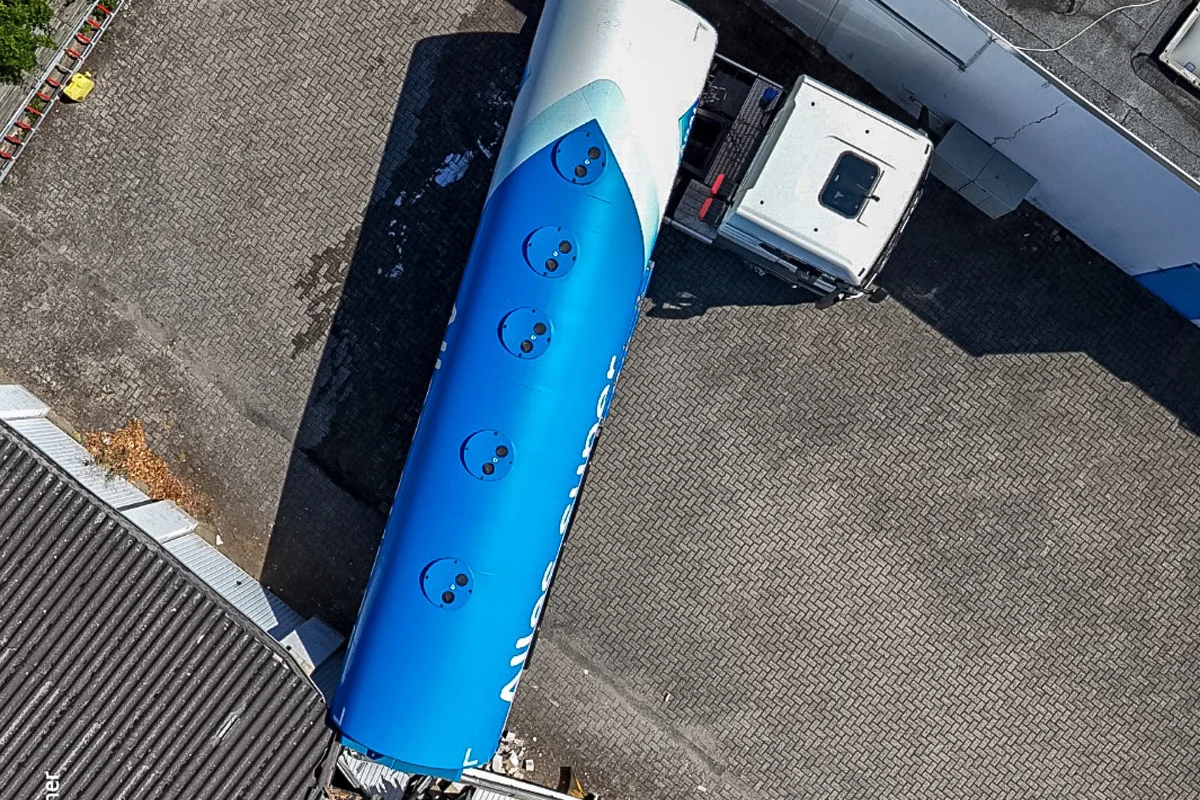 Ein LKW mit einem blauen Tankanhänger steht auf einem gepflasterten Hof neben einem Gebäude. Der Tank ist zylinderförmig und blau gefärbt.