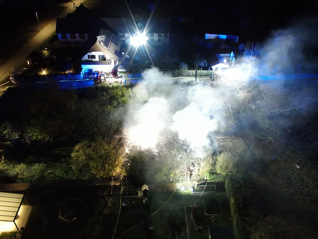 Bei Nacht ist ein Haus von oben zu sehen, das in Flammen steht. Dicker Rauch steigt auf, während Einsatzfahrzeuge mit blinkenden Lichtern vor Ort sind.