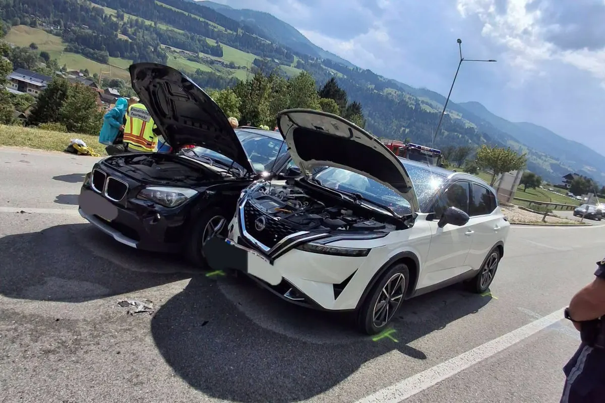Zwei Autos, ein schwarzer BMW und ein weißer Nissan, stehen mit geöffneten Motorhauben auf einer Straße nach einem Unfall. Im Hintergrund sind Rettungskräfte zu sehen.