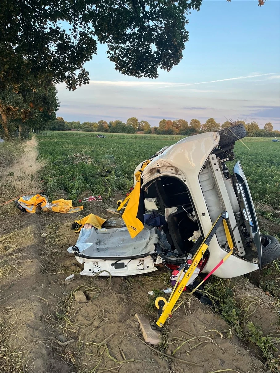 Ein weißes Auto liegt umgestürzt auf seiner Seite in einem Feld am Rand einer Baumreihe. Rettungswerkzeug ist erkennbar.