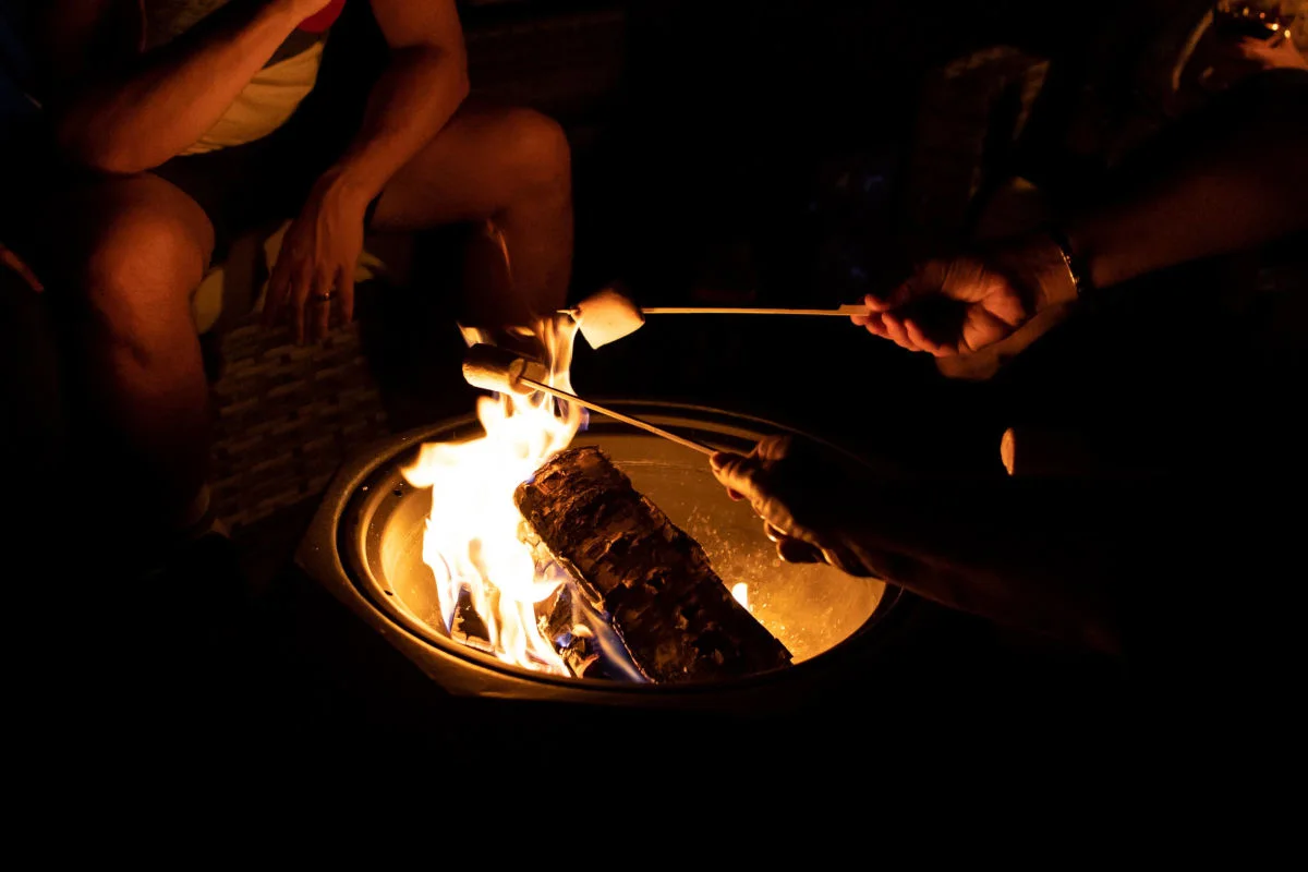 Zwei Personen halten Stöcke mit Marshmallows über einer Feuerschale, während das Feuer brennt. Eine weitere Person sitzt daneben.