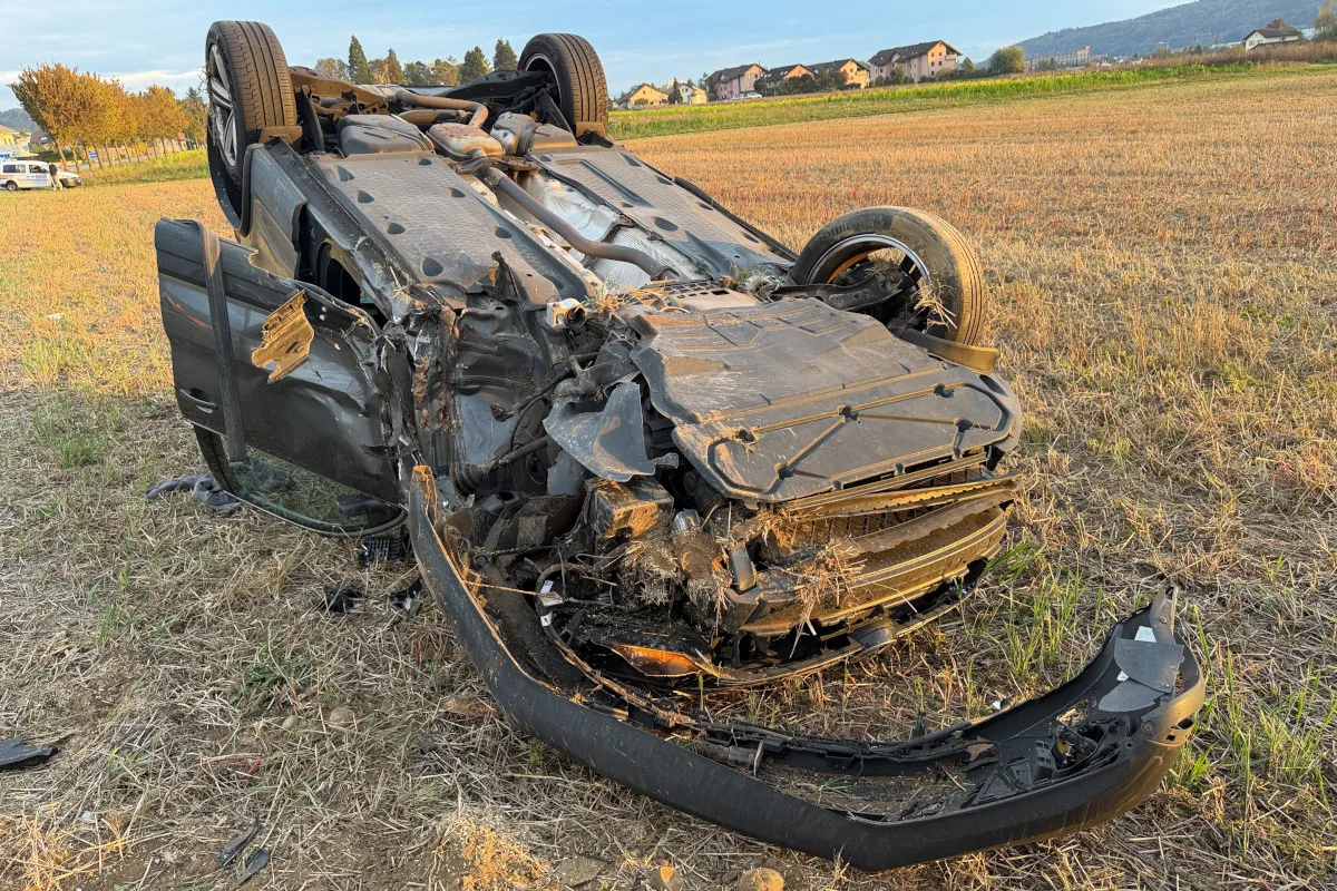 Ein Auto ist schwer beschädigt und liegt auf dem Dach auf einem Feld. Im Hintergrund sind Wohnhäuser zu sehen.