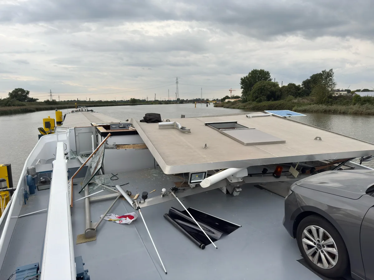 Ein Binnenschiff zeigt erheblichen Schaden auf seinem Deck, inklusive zerbrochenem Glas und herumliegenden Trümmern. Am Rand steht ein Auto.