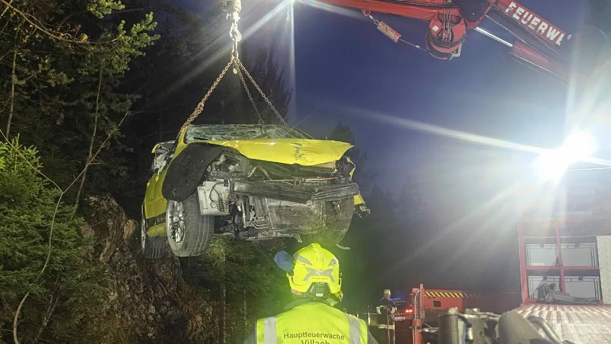 Ein gelbes Auto mit schweren Schäden wird nachts von einem Kran gehoben. Feuerwehrleute sind im Hintergrund zu sehen.