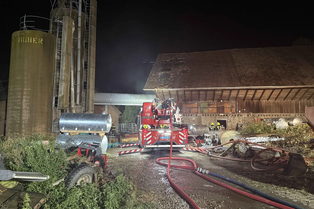 Ein Feuerwehrfahrzeug mit ausgefahrenen Schläuchen steht vor einer Scheune, die deutliche Brandschäden aufweist. Im Hintergrund sind Silos und Feuerwehrleute in Aktion zu sehen.