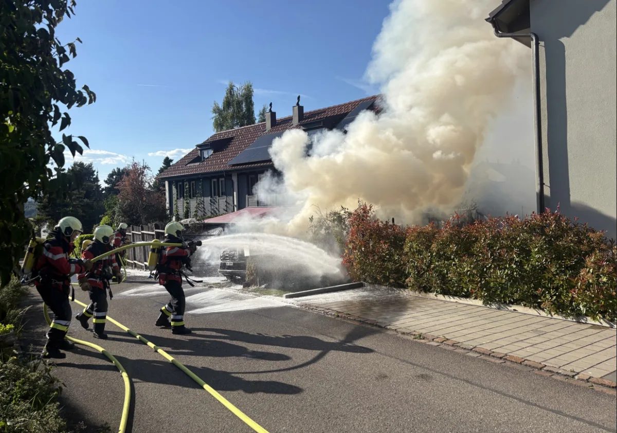 Mehrere Feuerwehrleute in Schutzanzügen löschen mit Wasserschläuchen ein großes Feuer, das dichten Rauch neben einem Wohnhaus produziert. Der Himmel ist klar und die Szene spielt sich in einem Vorort ab.