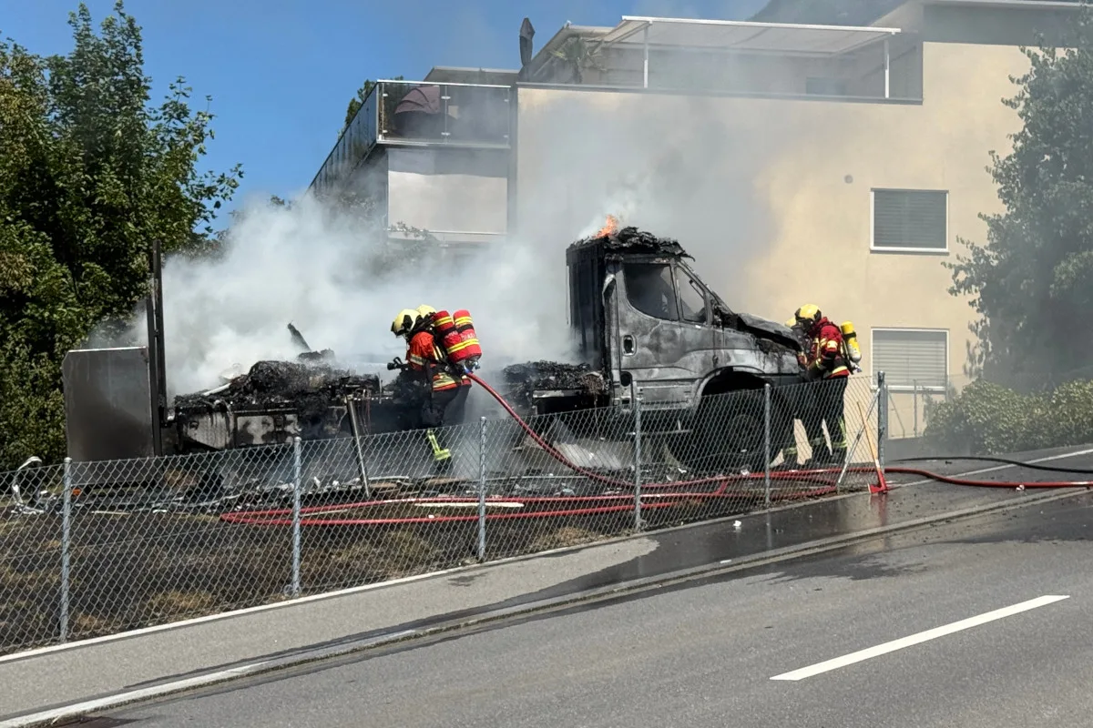 Zwei Feuerwehrleute versuchen, ein brennendes Fahrzeug mit Schläuchen zu löschen. Dicker Rauch steigt in die Luft.