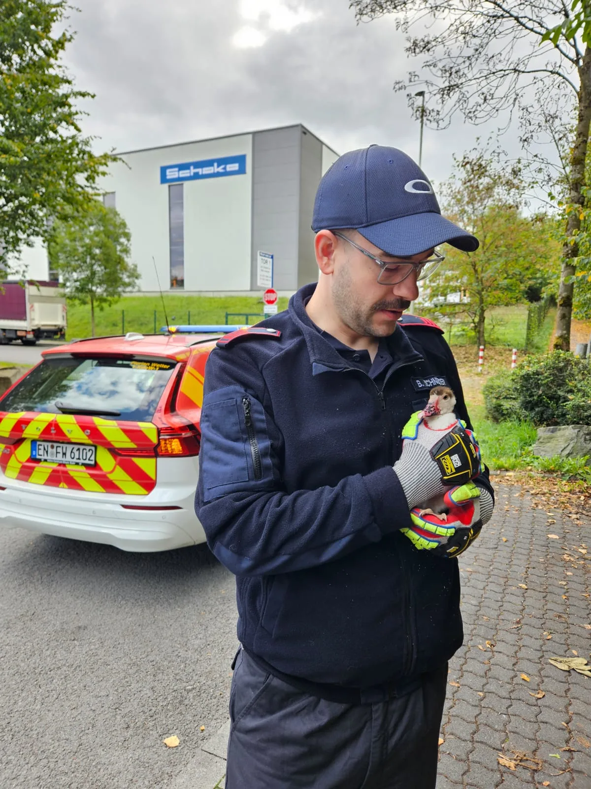 Ein Mann in dunkler Uniform mit Kappe hält behutsam ein kleines Tier in seinen Händen. Im Hintergrund steht ein rot-gelb markiertes Einsatzfahrzeug und ein Industriegebäude.