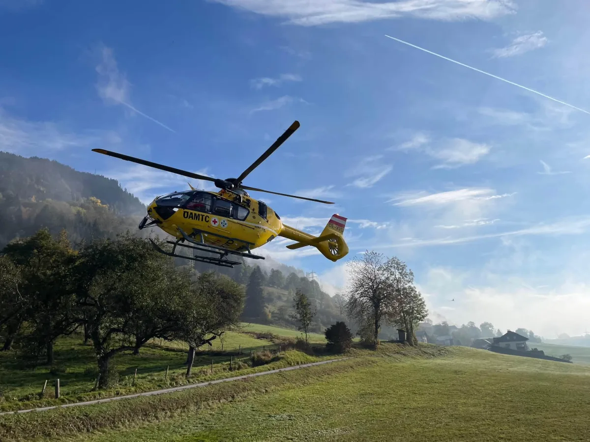Ein gelber Rettungshubschrauber fliegt knapp über einer grünen Wiese, im Hintergrund sind Bäume und Hügel.