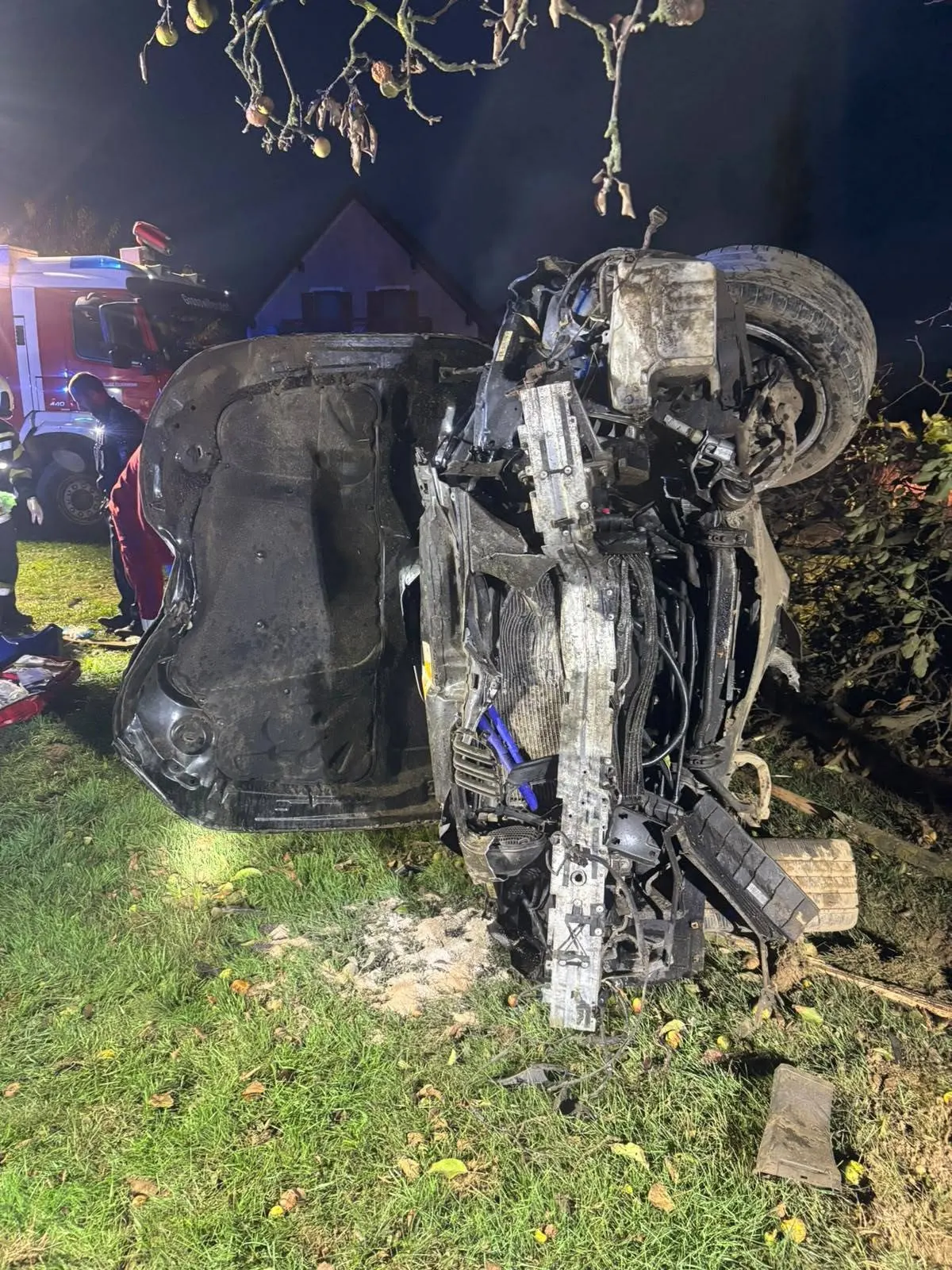 Ein Auto liegt auf der Seite in einer Grünfläche, umgeben von Feuerwehrleuten und einem Feuerwehrfahrzeug im Hintergrund. Äste hängen im Vordergrund herunter.